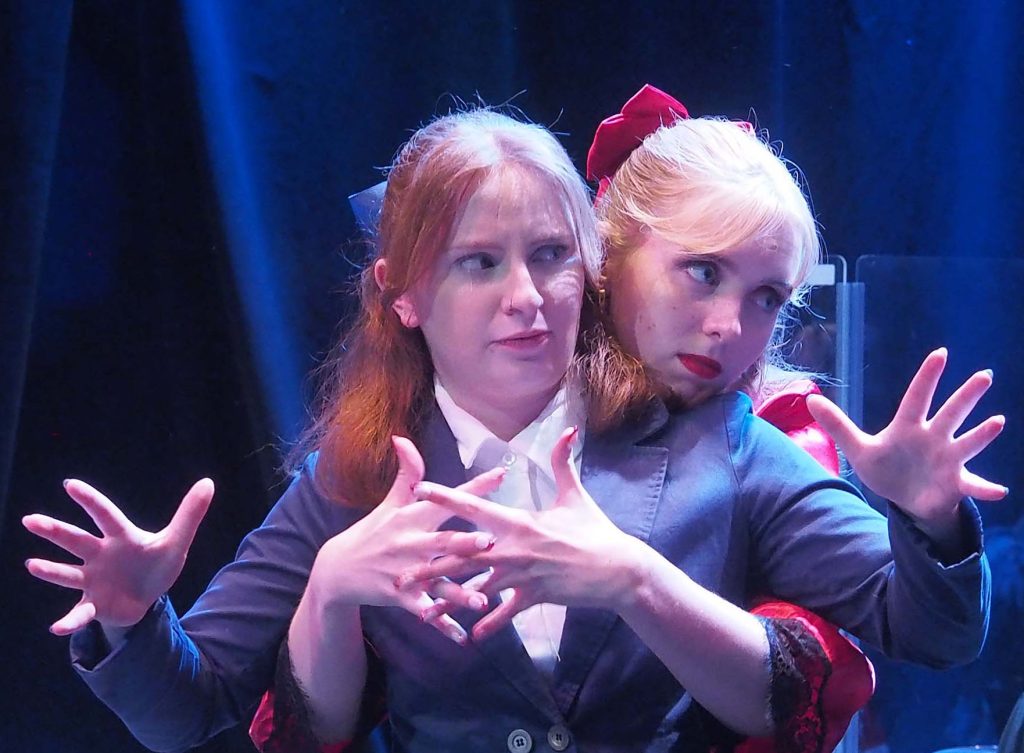 Two performers face out while one is behind the other and laces her fingers across the front performer's chest.