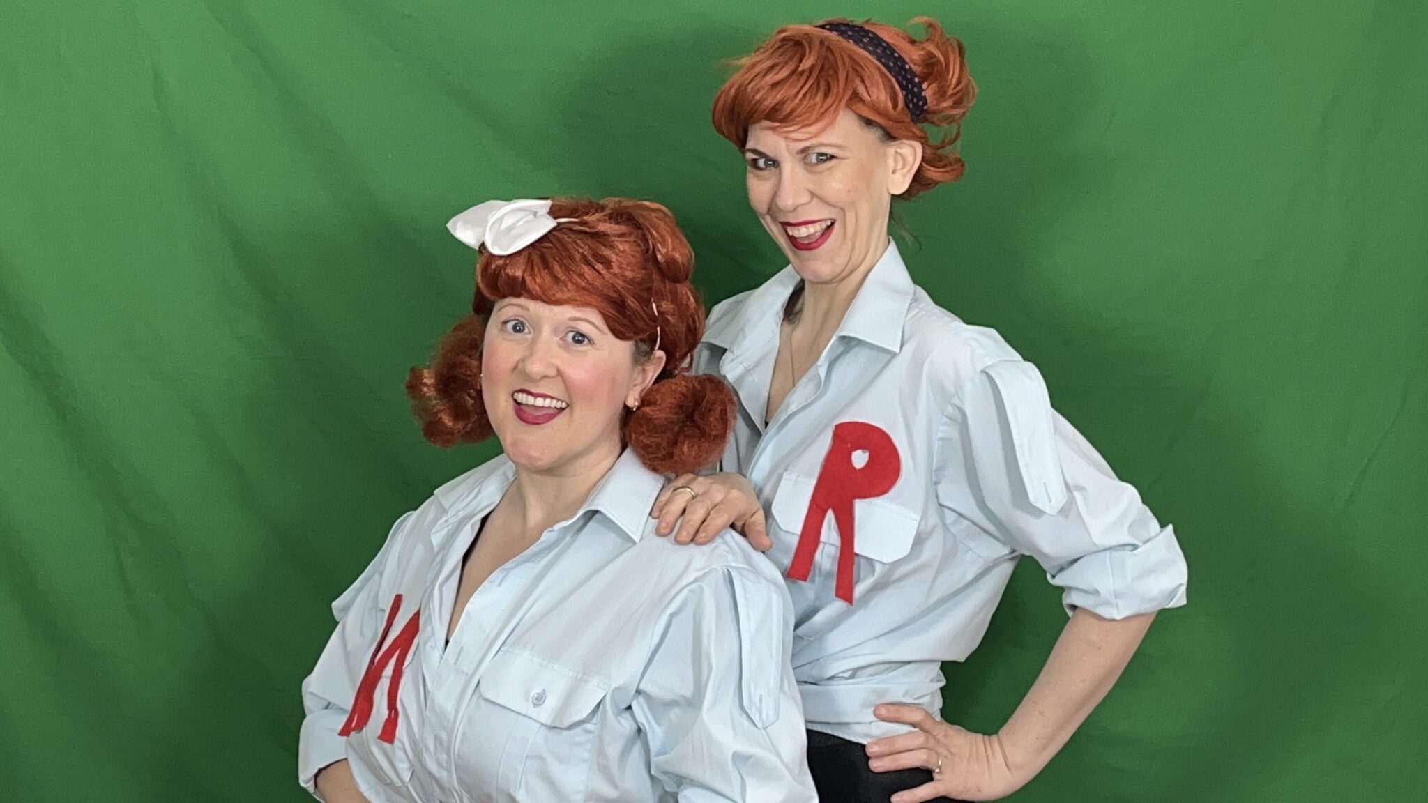 Two performers wearing collared shirts, both with red hair, pose in front of a green screen.