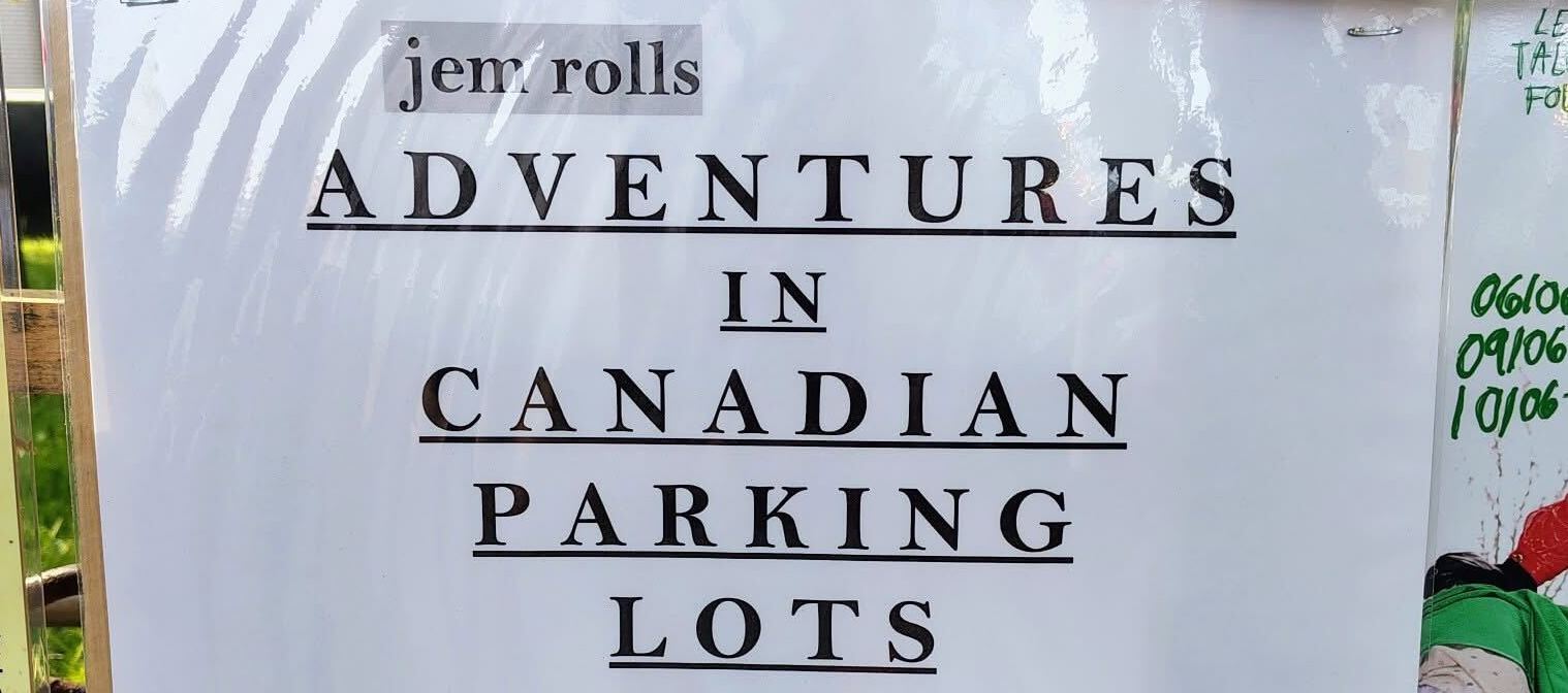 a white paper sign with black text reading: "ADVENTURES IN CANADIAN PARKING LOTS"