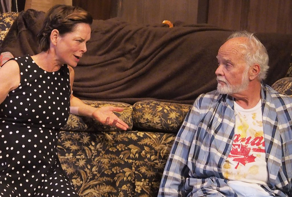 Two people sitting against a couch, speaking.