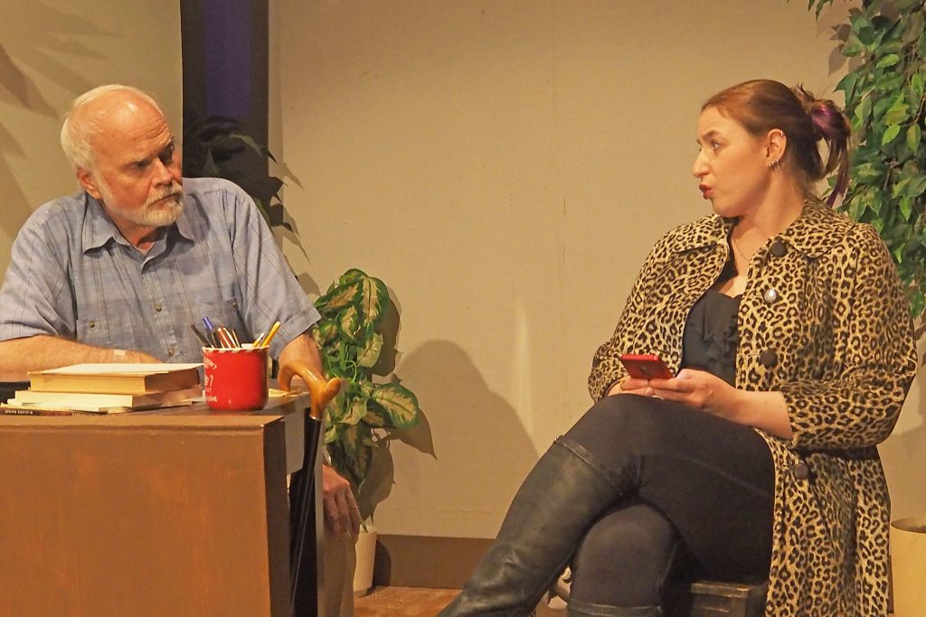 One person sits at a desk with books, the other in a chair beside the desk. They speak.