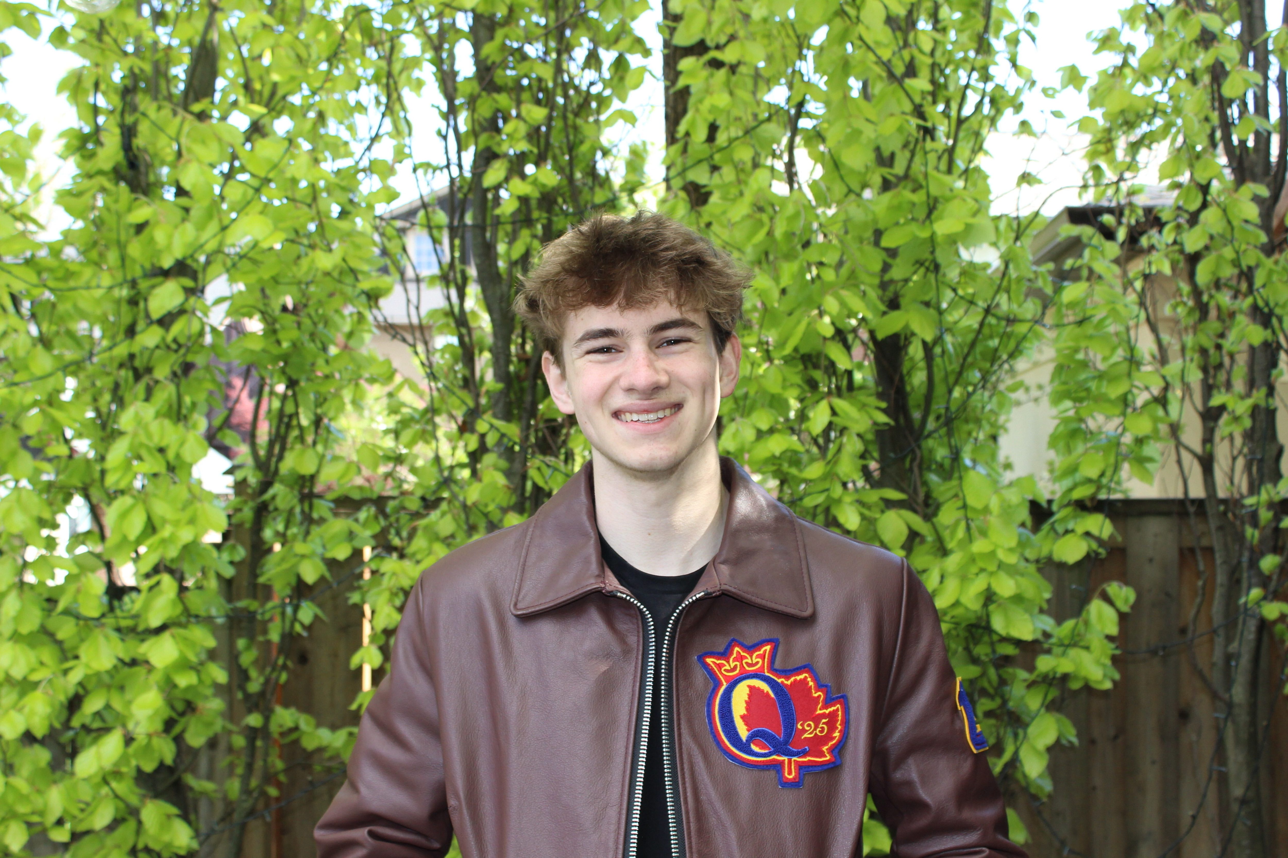 A white man with brown hair wearing a brown leather jacket, smiling in front of a backdrop of trees.