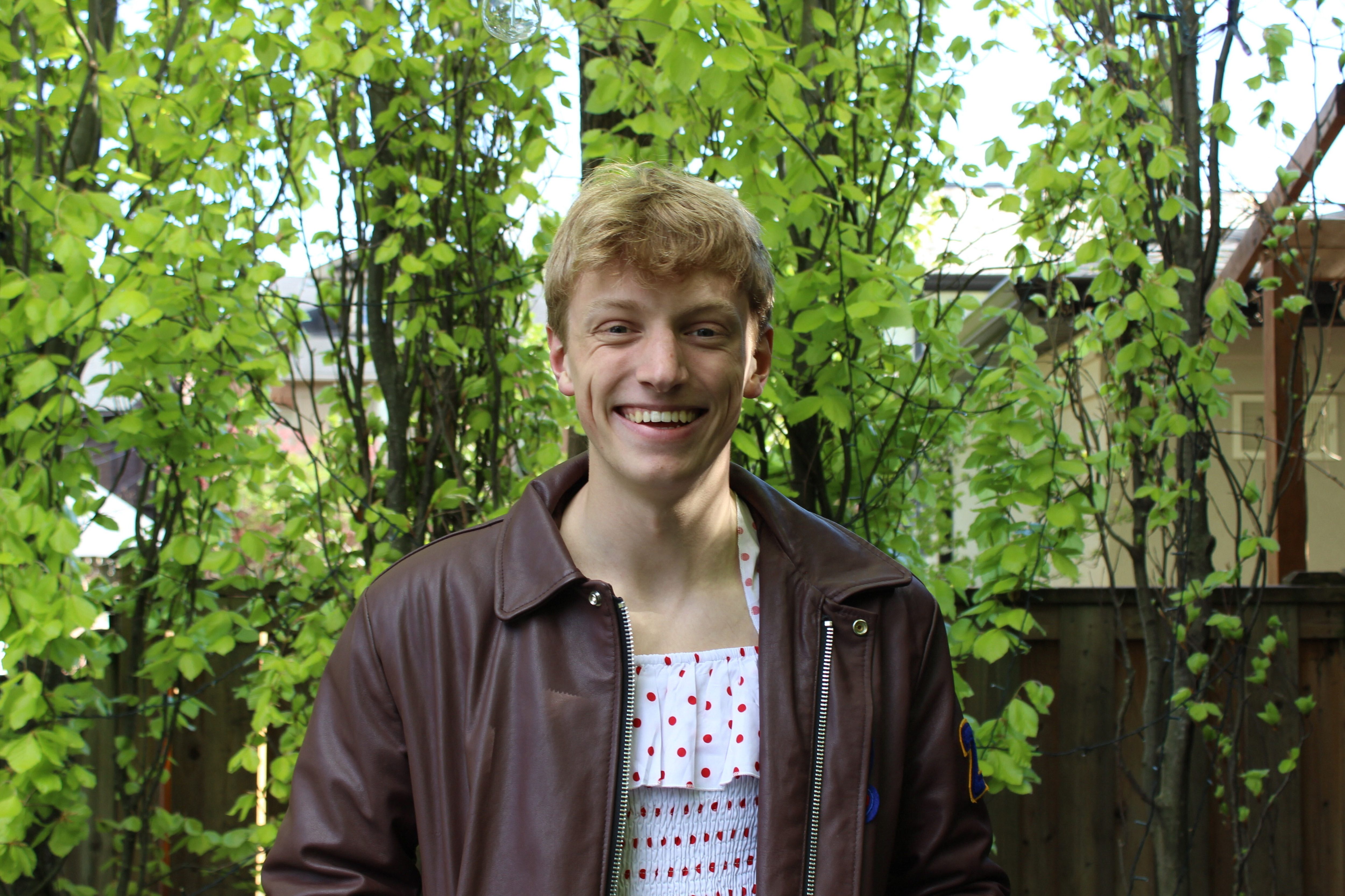 A blonde man in a brown leather jacket smiling in front of a backdrop of trees.