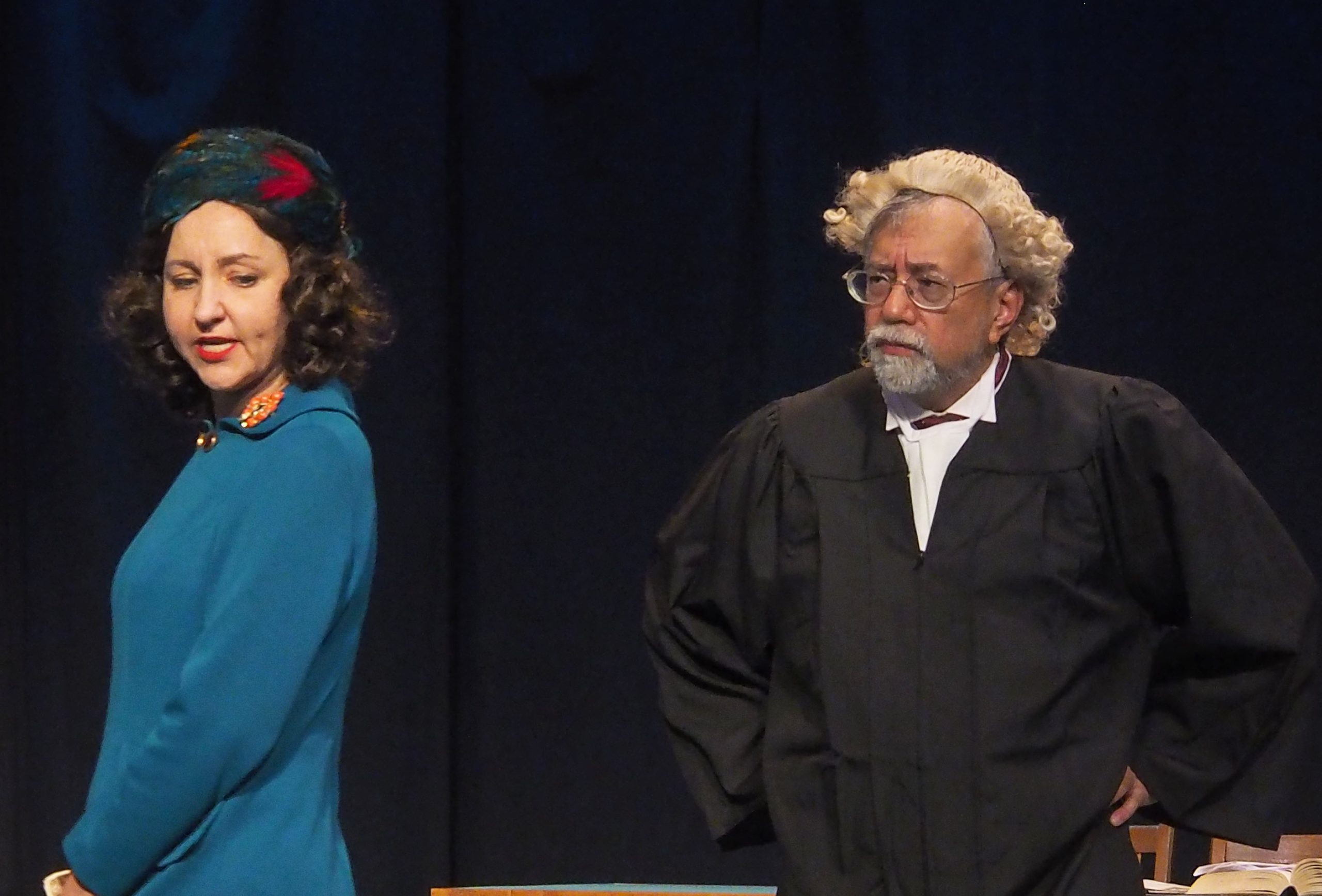 Two performers in Domino Theatre's production of 'Witness for the Prosecution'. One actor wears a blue coat and looks over her shoulder. The other actor is dressed as a judge and looks concernedly at the other actor.