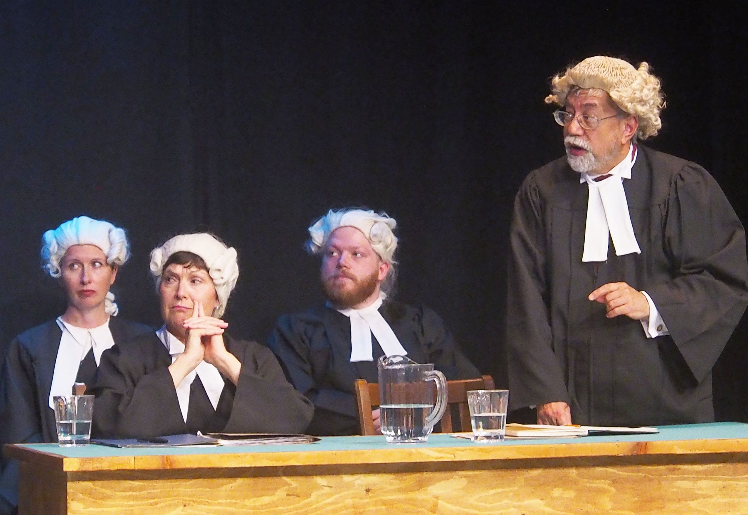 Four performers in Domino Theatre's production of 'Witness for the Prosecution'. One stands to speak and the other three sit.