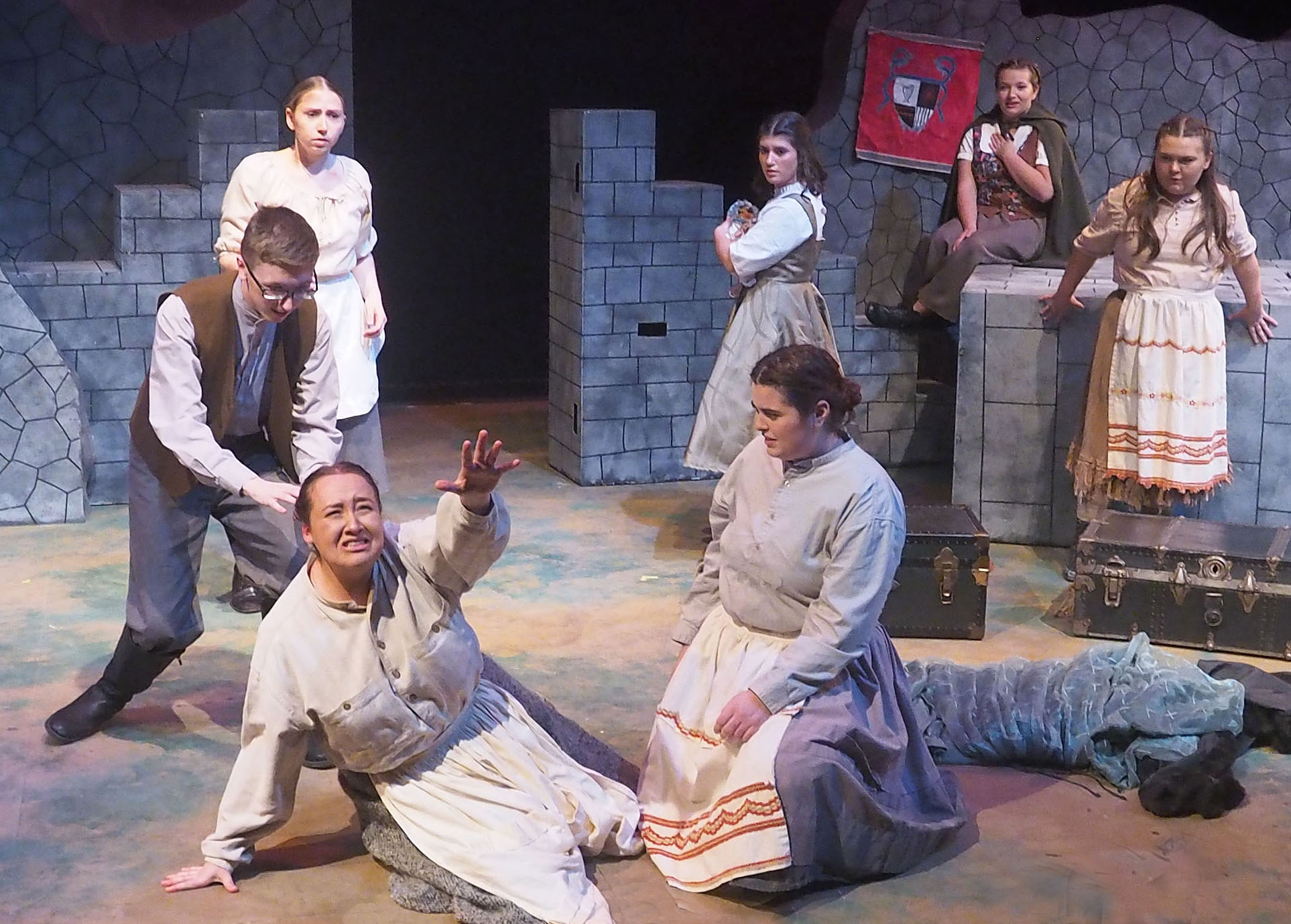 Actors performing on a stage. The setting is stone brick walls and various actors wear drab dresses, pants, and shirts. The forefront actor looks very distressed and is reaching upwards. 