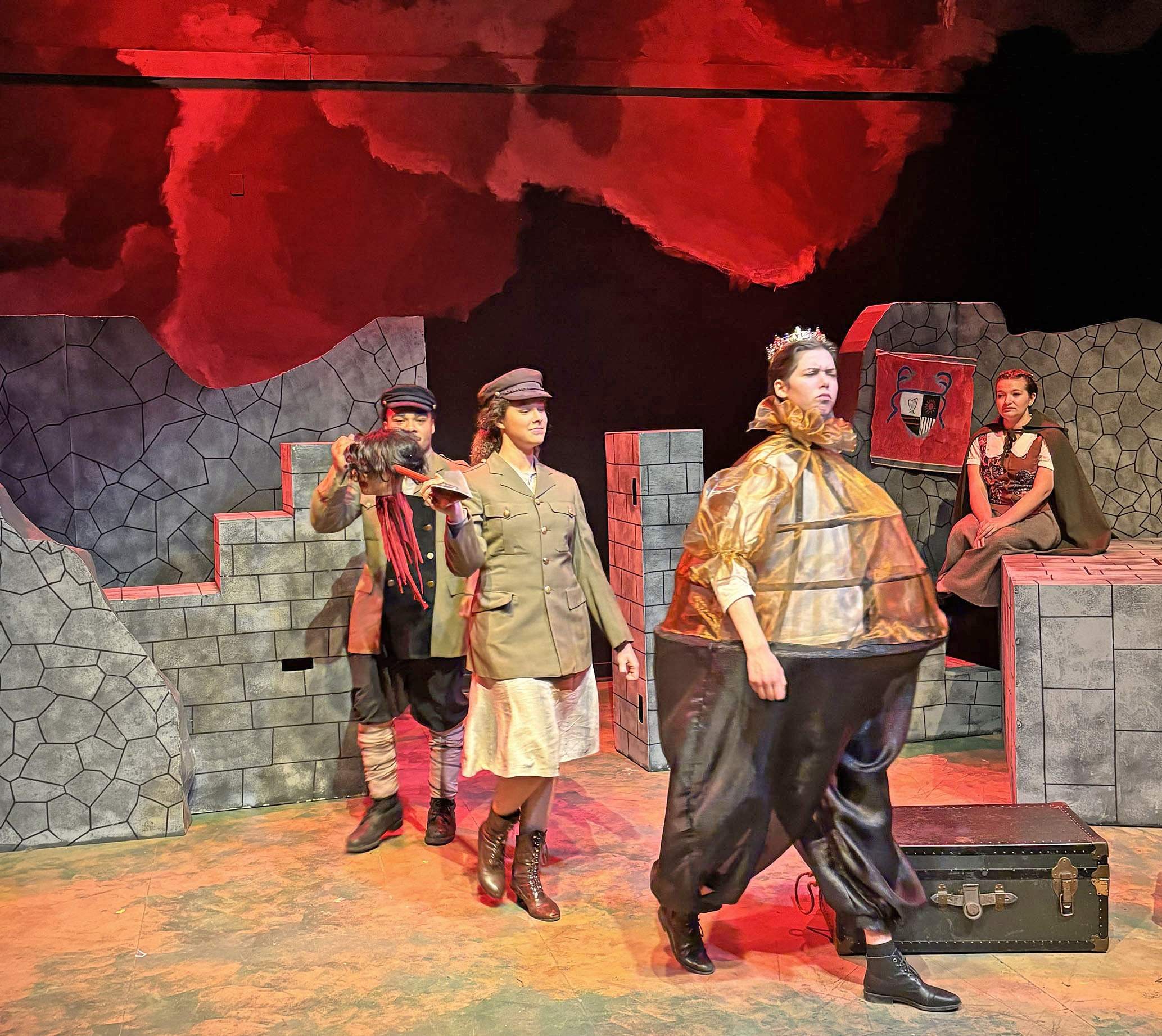 Actors on a stage. The setting is stone brick walls. The forefront actor is dressed very regally in a large suit and crown.