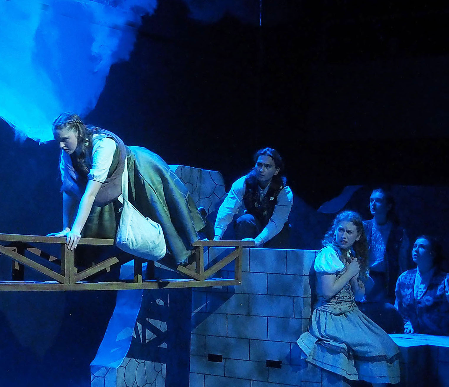 Actors performing on a stage. One actor is crossing a beam while the others watch nervously. 