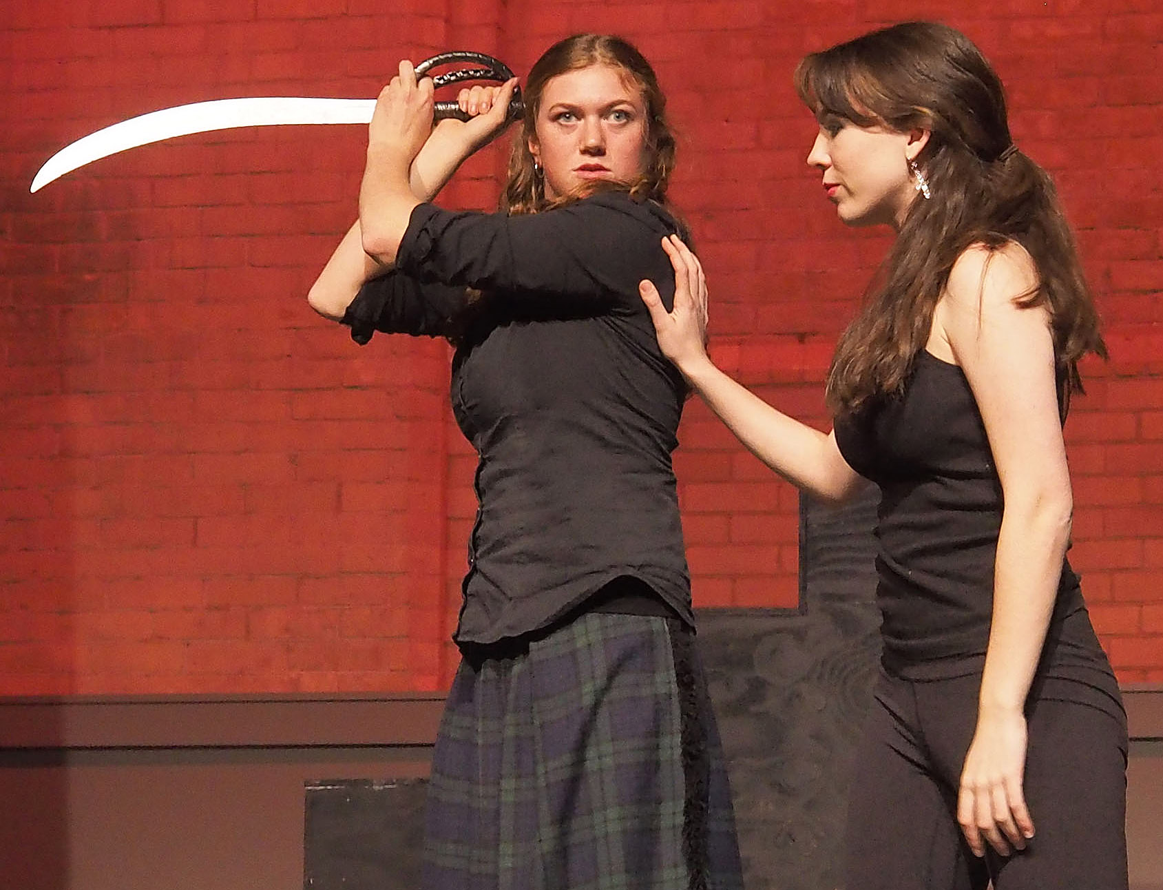 Two actors performing on stage. One holds a sword and looks straight ahead. The other has their hand pressed against the sword-wielding actor. 