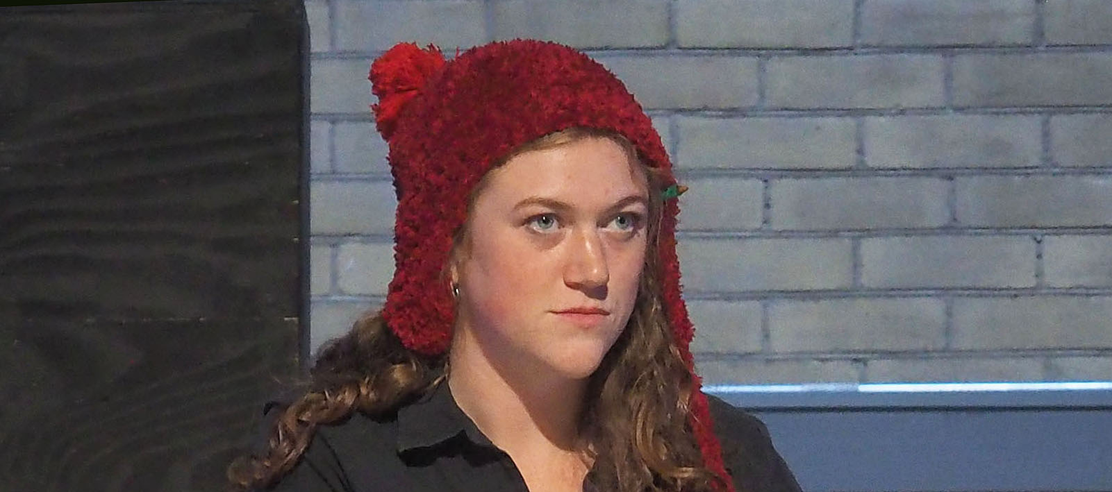 actor sits on stage behind a desk. She wears a black top and red winter hat while looking sternly.