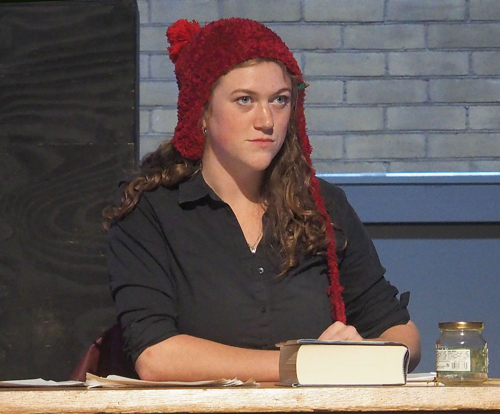 An actor performing on stage. They sit at a desk and stare straight ahead. 