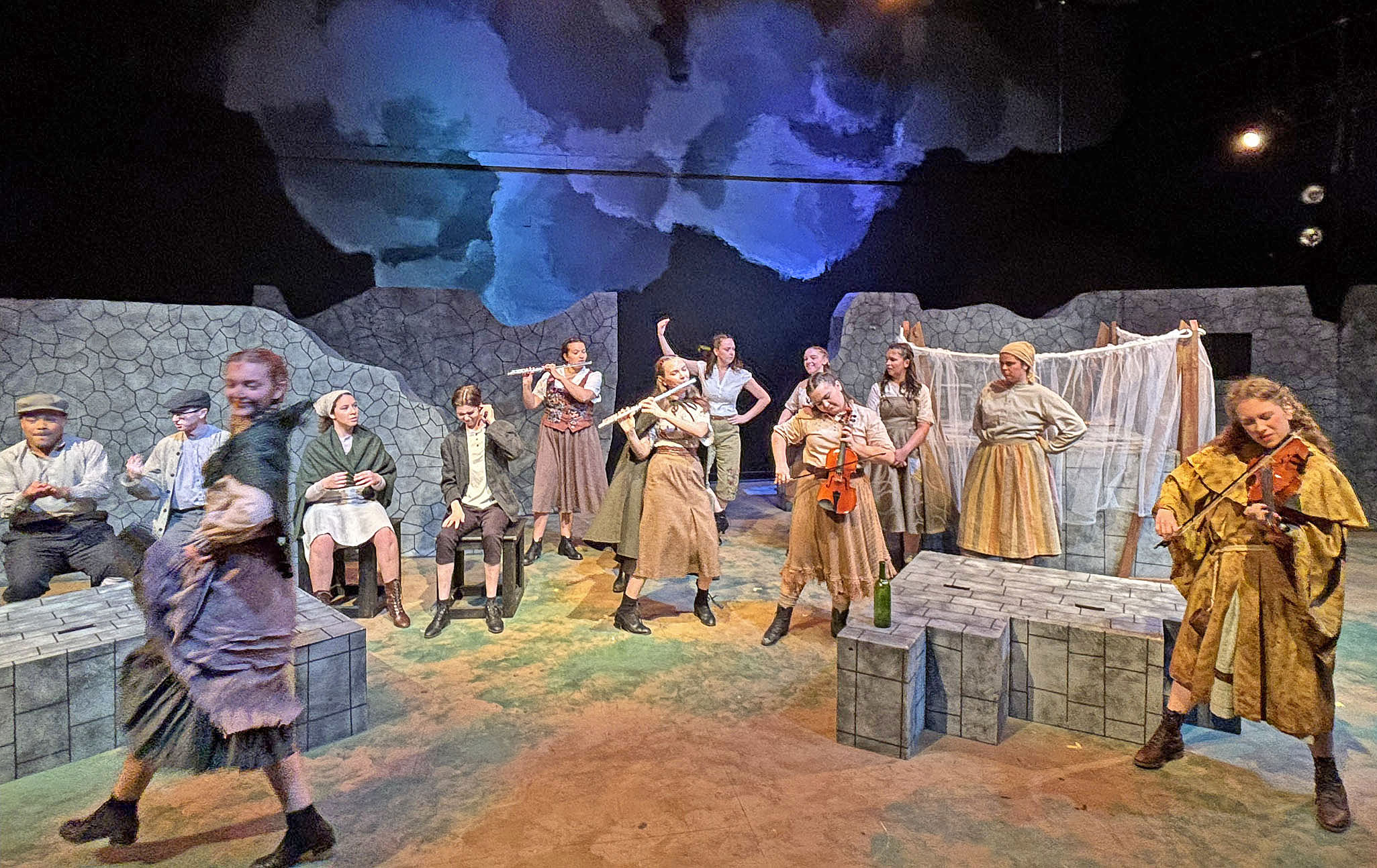 Many actors on a stage, most of who play various instruments. Some appear to be dancing. The setting is stone brick and the actors wear old-fashioned, drab attire. 