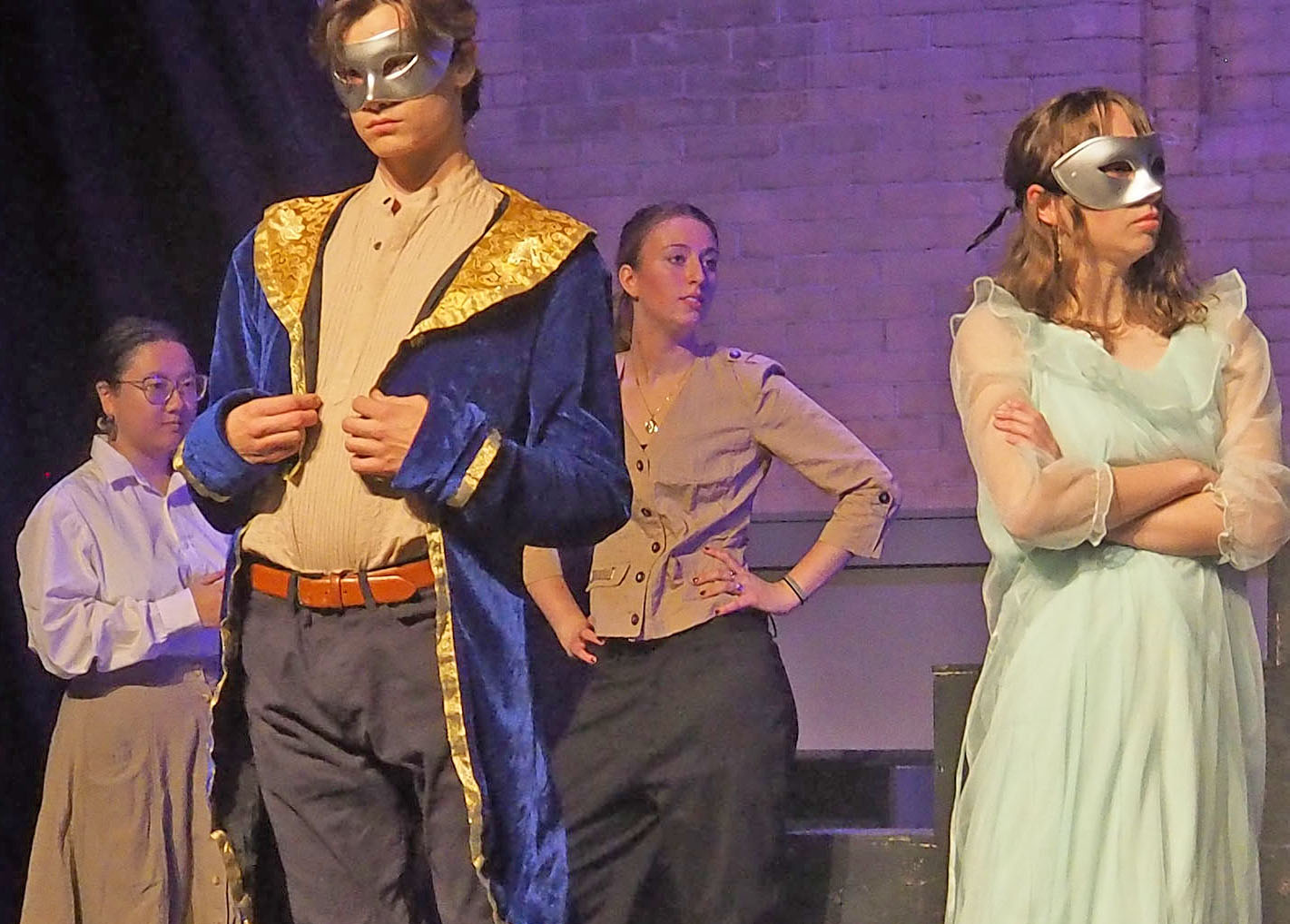 Four actors performing on a stage. The lighting has a purple hue and two of the actors wear silver masks. One actor wearing a mask looks bothered while the other masked actor appears to show off their coat. The two unmasked actors look off to the side. 