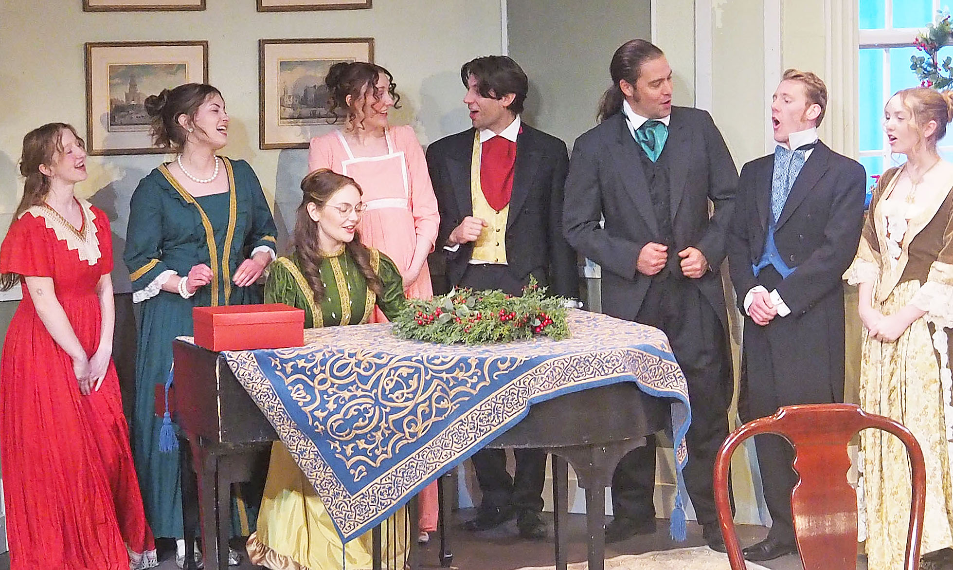 A group of eight actors performing. The setting is old fashioned and the actors are dressed in dresses and suits of 17th century style. One actor sits at a piano, playing it, and the others stand behind her, appearing to sing. 