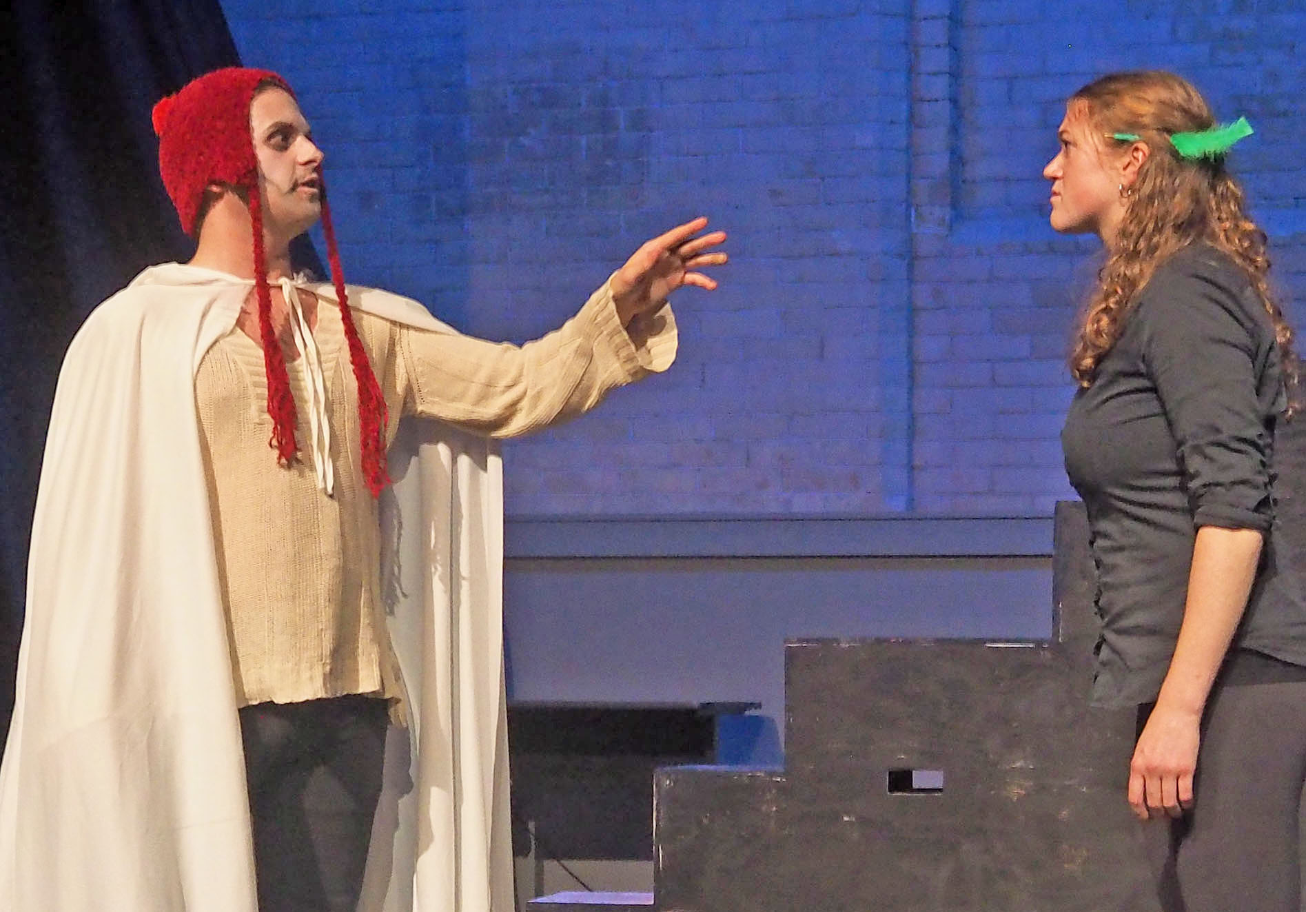 Two actors performing on stage. They look at each other. One reaches out to the other wearing a cape and has a ghoulish expression. The other stares back with a feather in their hair. 