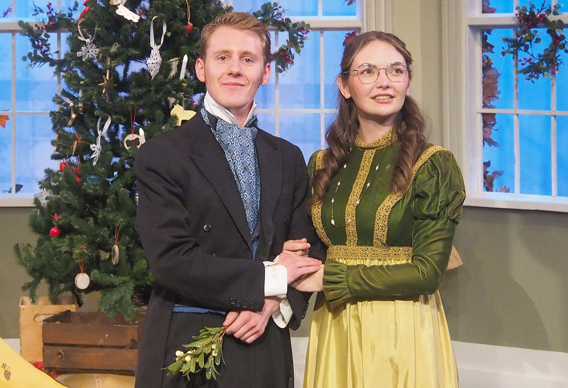 Two actors stand in front of a Christmas tree, appearing to pose for a photo. The hold hands and are both wearing styles of the 17th century. 