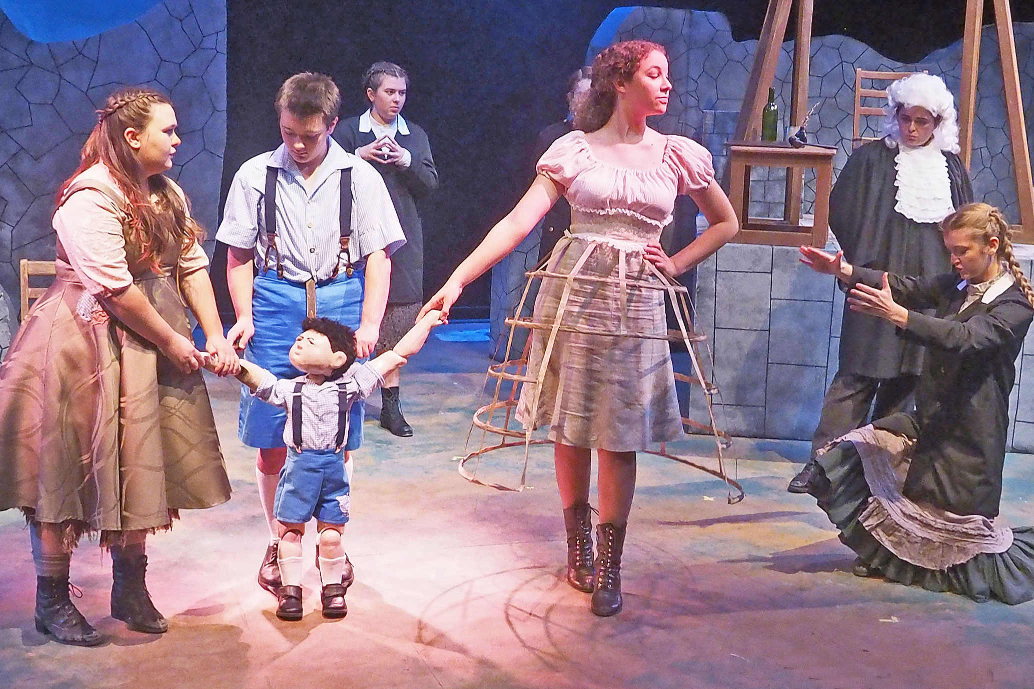 Actors on a stage. The setting is stone brick walls. A few actors hold onto a puppet. Most actors are dressed in drab clothing while one is dressed as an old-fashioned judge. 