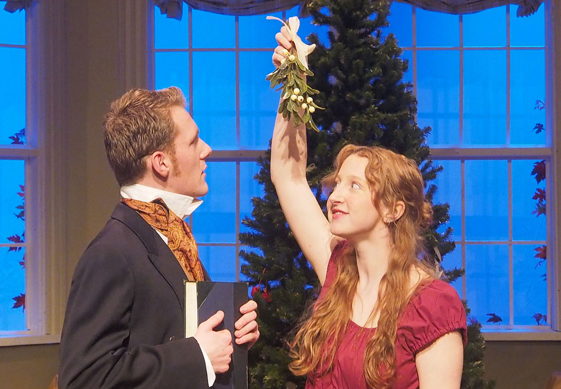 Two actors stand below mistletoe in front of a Christmas tree. 