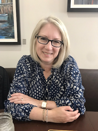 Headshot of a woman with blonde hair. She is sitting and leans against a table. She smiles.