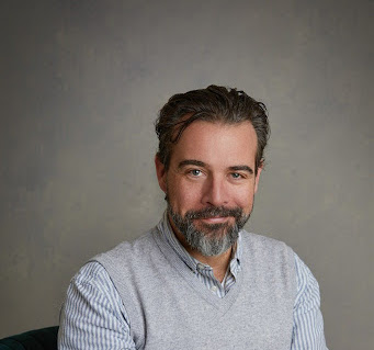 Headshot of a man against a grey background. He wears a lightly coloured collared shirt.