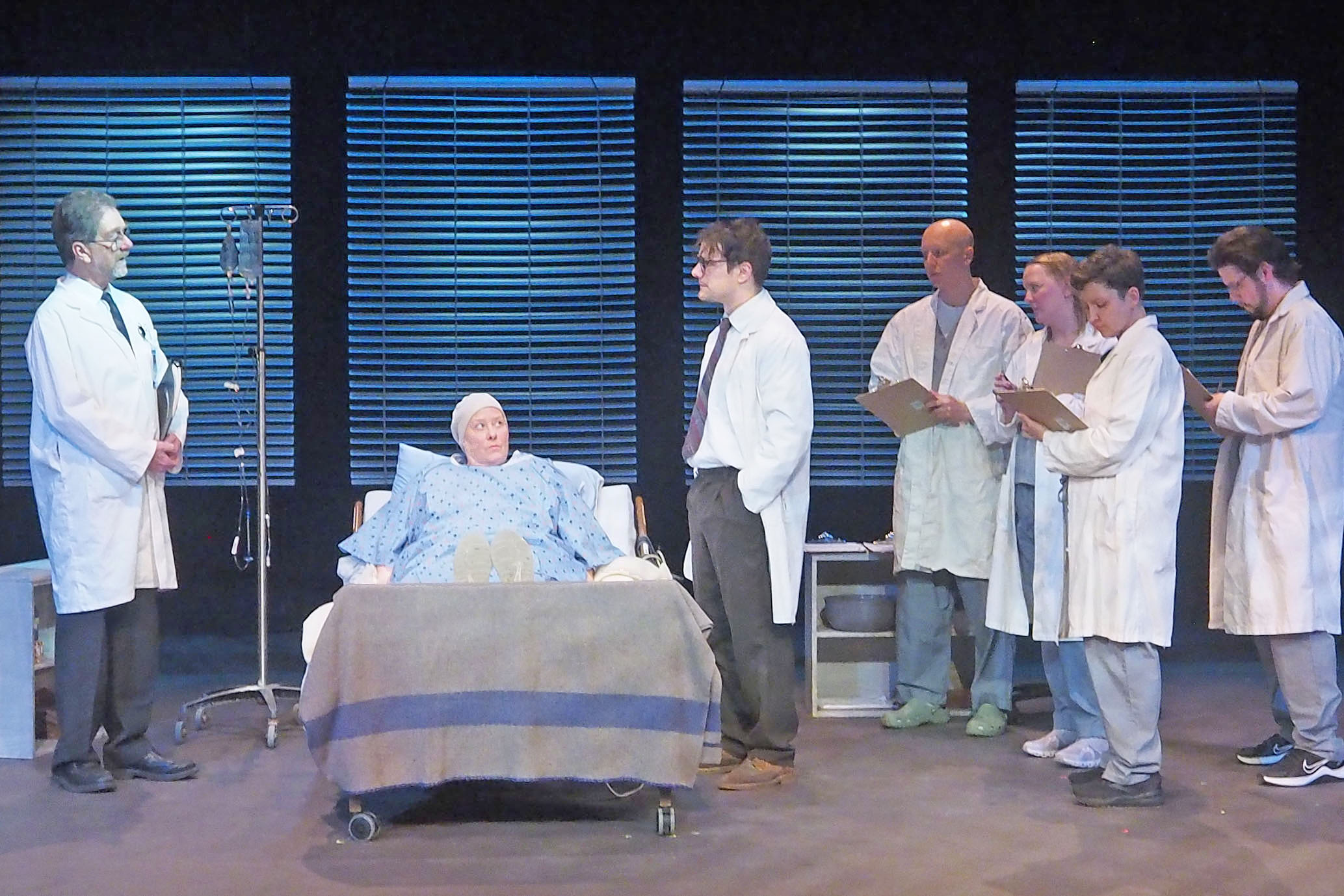 A stage performance where a hospital patient lies in a hospital bed. They are speaking to a set of doctors and nurses who are either listening or writing things down. 