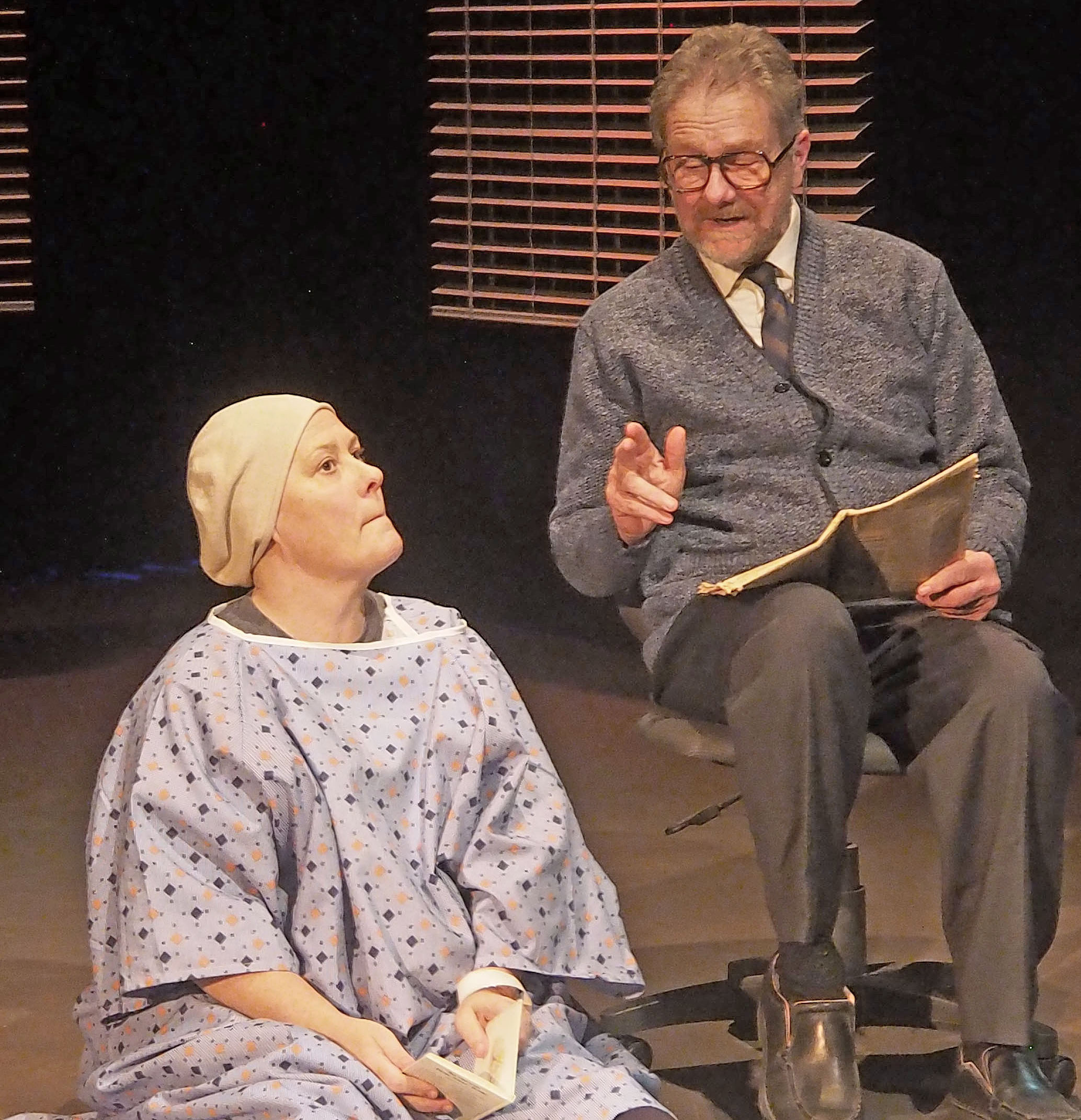 A stage performance where a hospital patient sits on the ground speaking to someone dressed in a tie, cardigan, and slacks. This individual sits on a chair holding a book. 