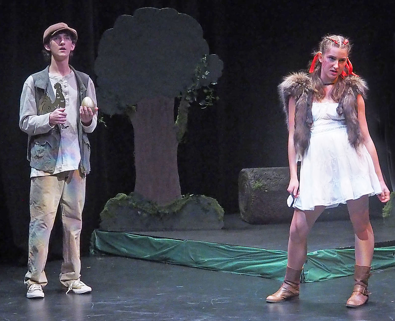 Two on stage performers stand in front of a stage tree. One wears a vest and holds a golden egg. The other actor wears a fur vest and has red bows in their hair. 