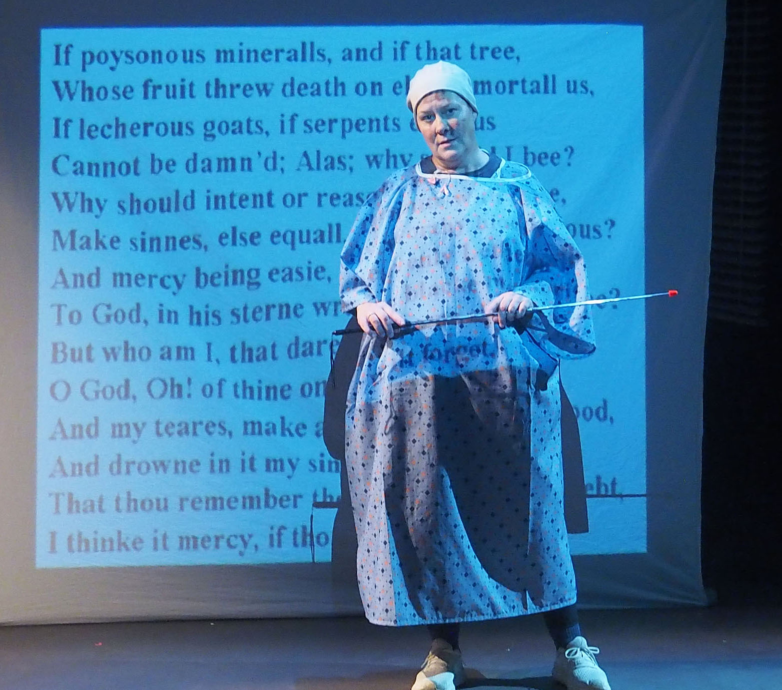 A stage performance where a hospital patient gives a presentation. They have a screen of words behind them and hold a pointer stick. 