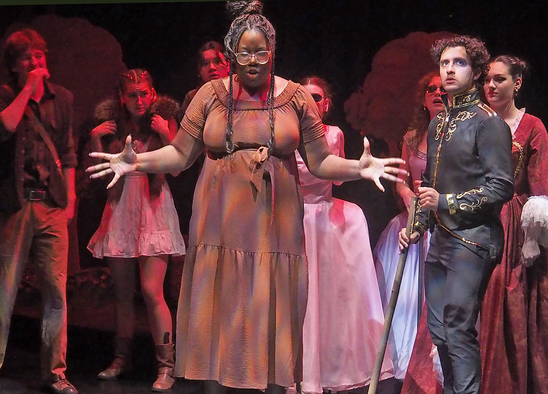 A group of performers stare at one performer front and centre. This performer wears a brown dress and has their arms partially stretched out. Another performer front and centre wears royal dresswear. 