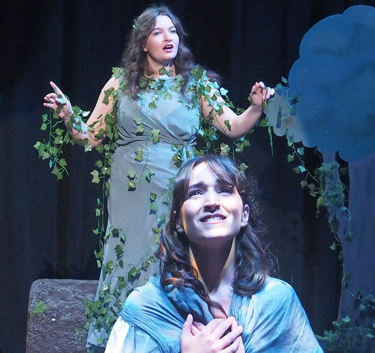 Two performers on stage. One is looking upwards. The performer behind them is dress as a tree.