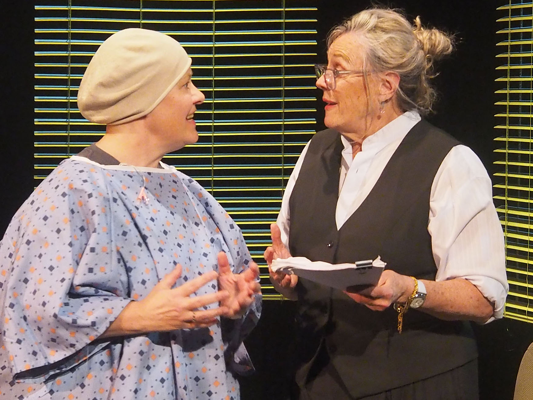 A stage performance where a hospital patient speaks to someone dressed in a collared shirt and vest, holding a stack of papers. 