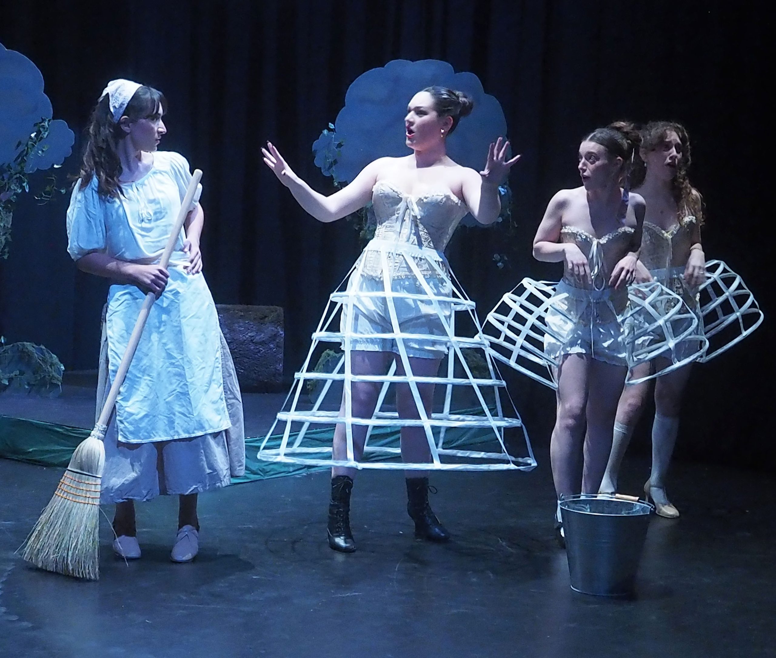 Performers on stage. One actor wears a an apron and carries a broom. The other three actors wear cage hoop skirts.
