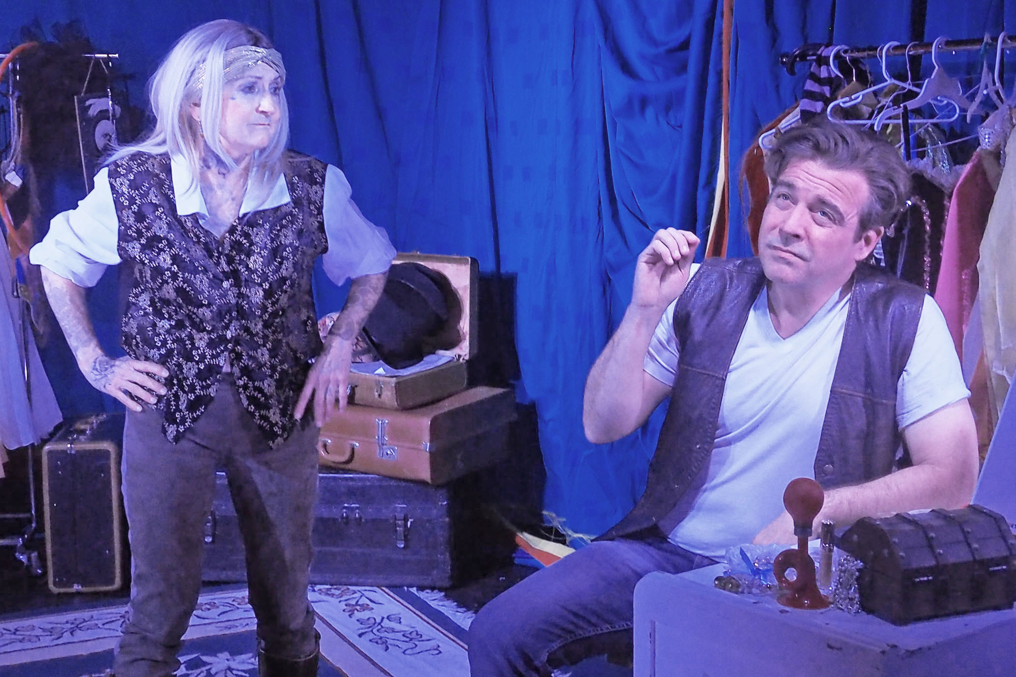 A man sits down, looking into the distance. A woman holds her hands on her hips while looking at him, seeming unimpressed. The background is a curtain and suitcases.