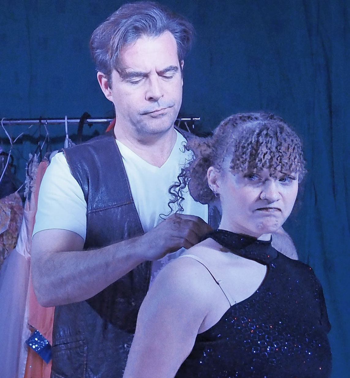 A man wear a vest helps to clip the neck of a woman's top. 