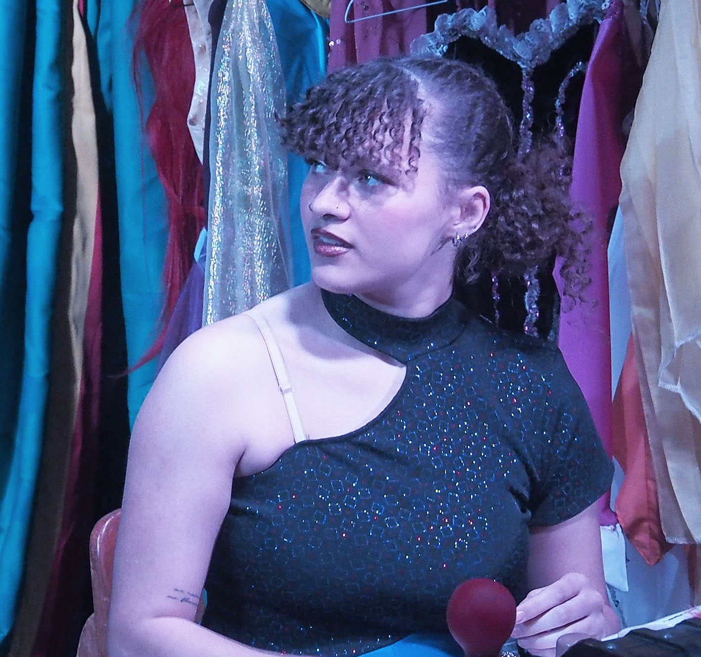 Woman wearing a one-shoulder black top looks off to the side. A clothing rack is in the background.