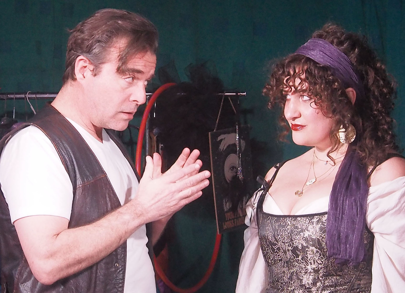 Two people speaking. One wears a vest and has his hands lifted to his chest. The other wears a headband that trails down her hair and a corset.