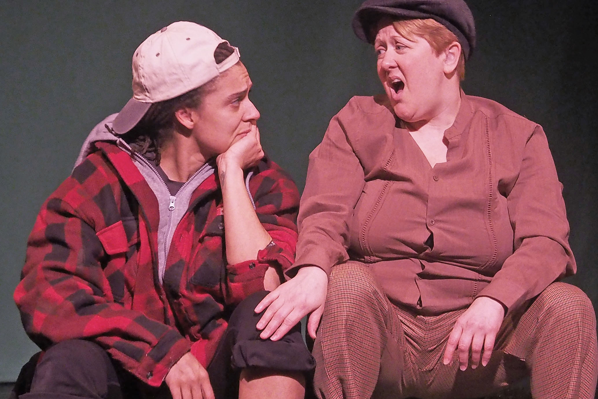 Two performers sit on stage. One looks at the other concernedly. The second performer appears distressed. 