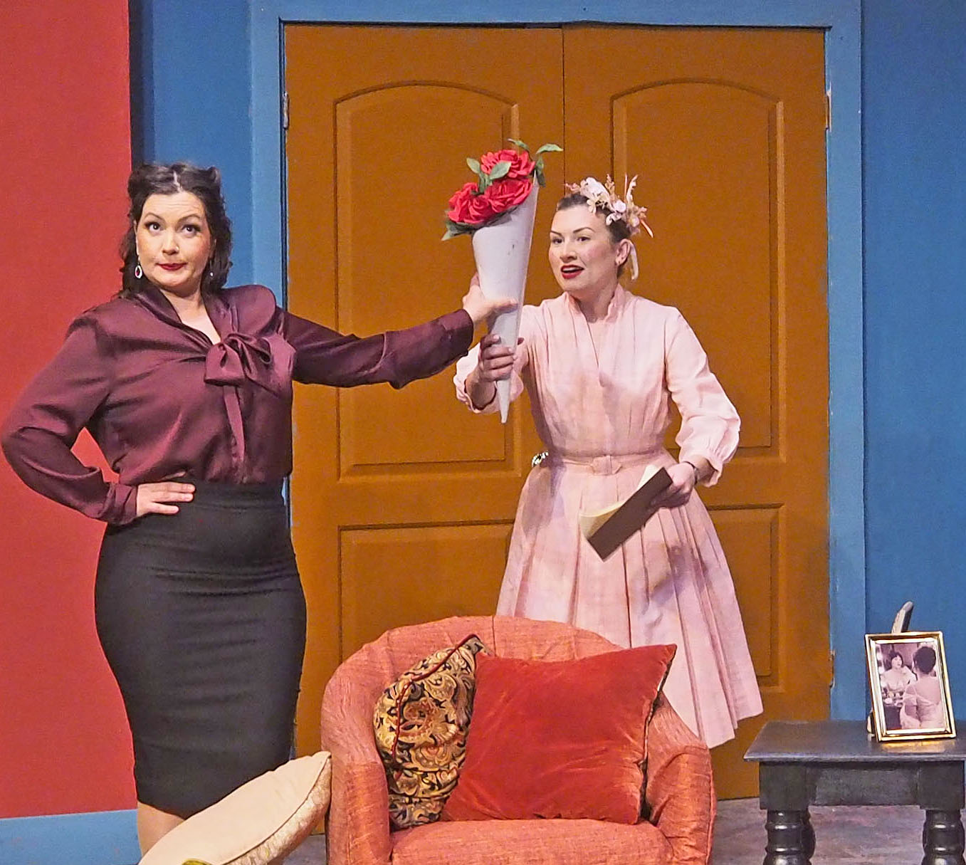 Two women are dressed elegantly. One looks away while handing the other a bouquet of roses.