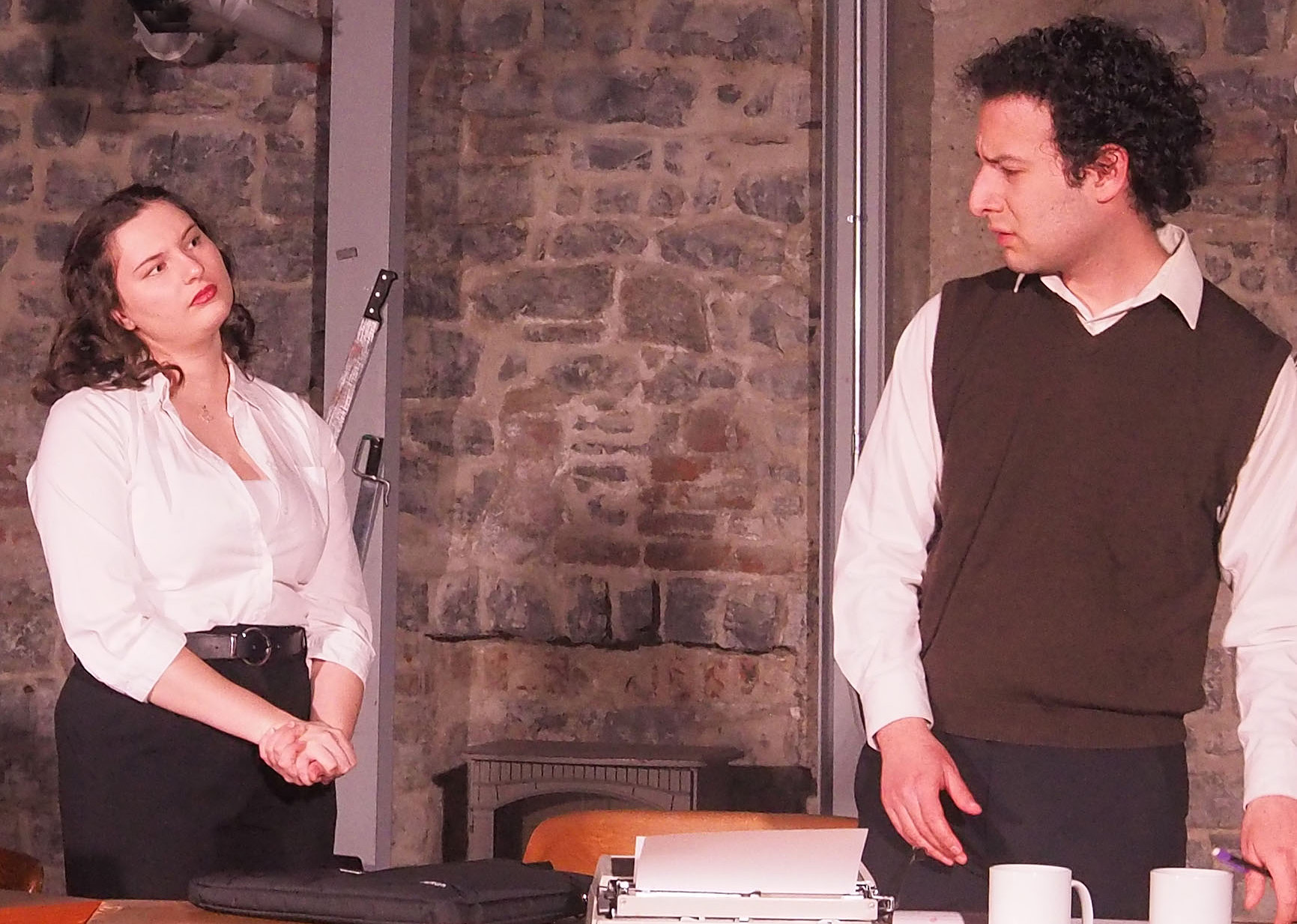 Two performers against a brick/stone wall backdrop. One performer looks concerned at the other while she looks confidently curious at him. There are two knives attached to the brick wall.