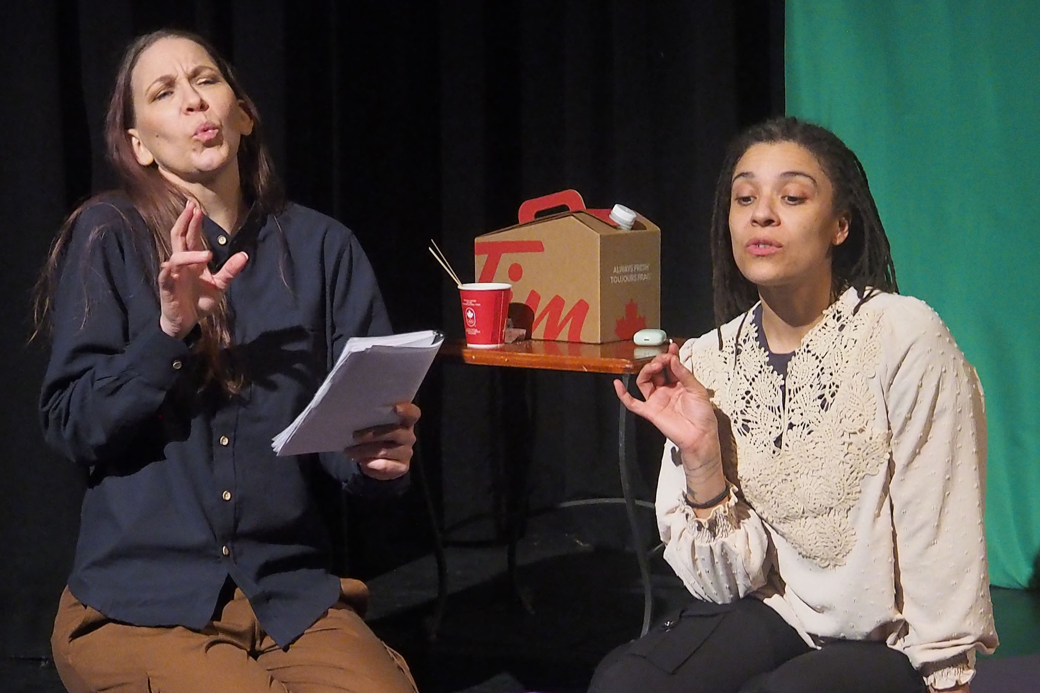 Two performers on stage, both sitting. One holds a stack of papers. There is a Tim Horton's coffee box and cup behind them. 