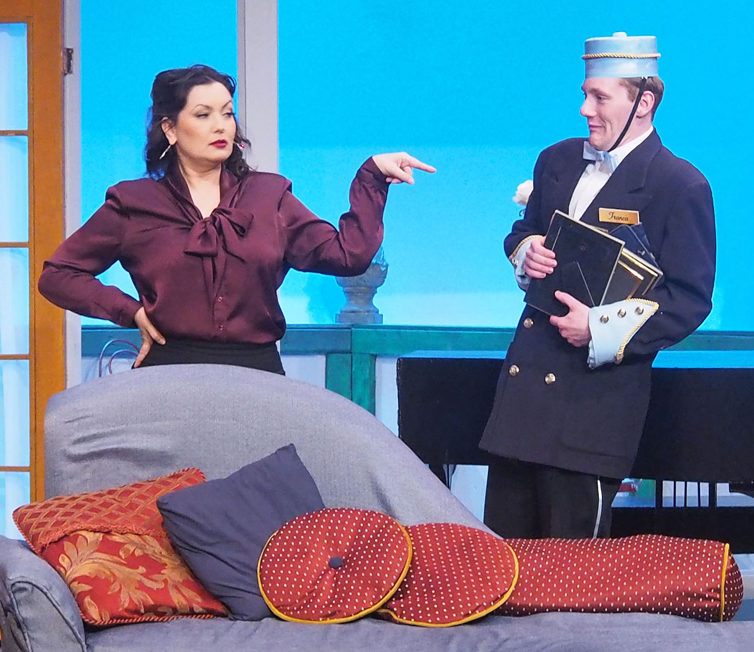 A woman dressed in high-end fashion points at a man dressed as a lobby boy. He holds picture frames and appears scared. They stand behind a couch decorated in pillows.