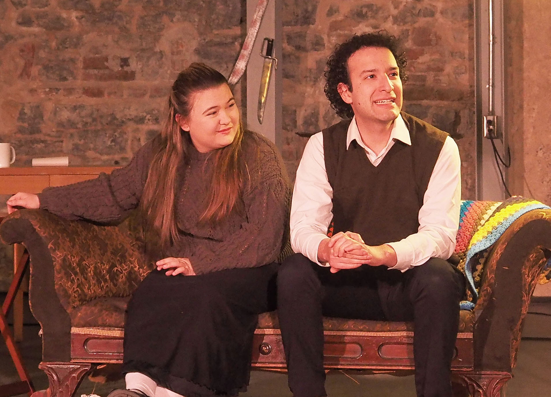 Two performers sitting on a brown couch against a beige-brown backdrop. They both look up at something. 