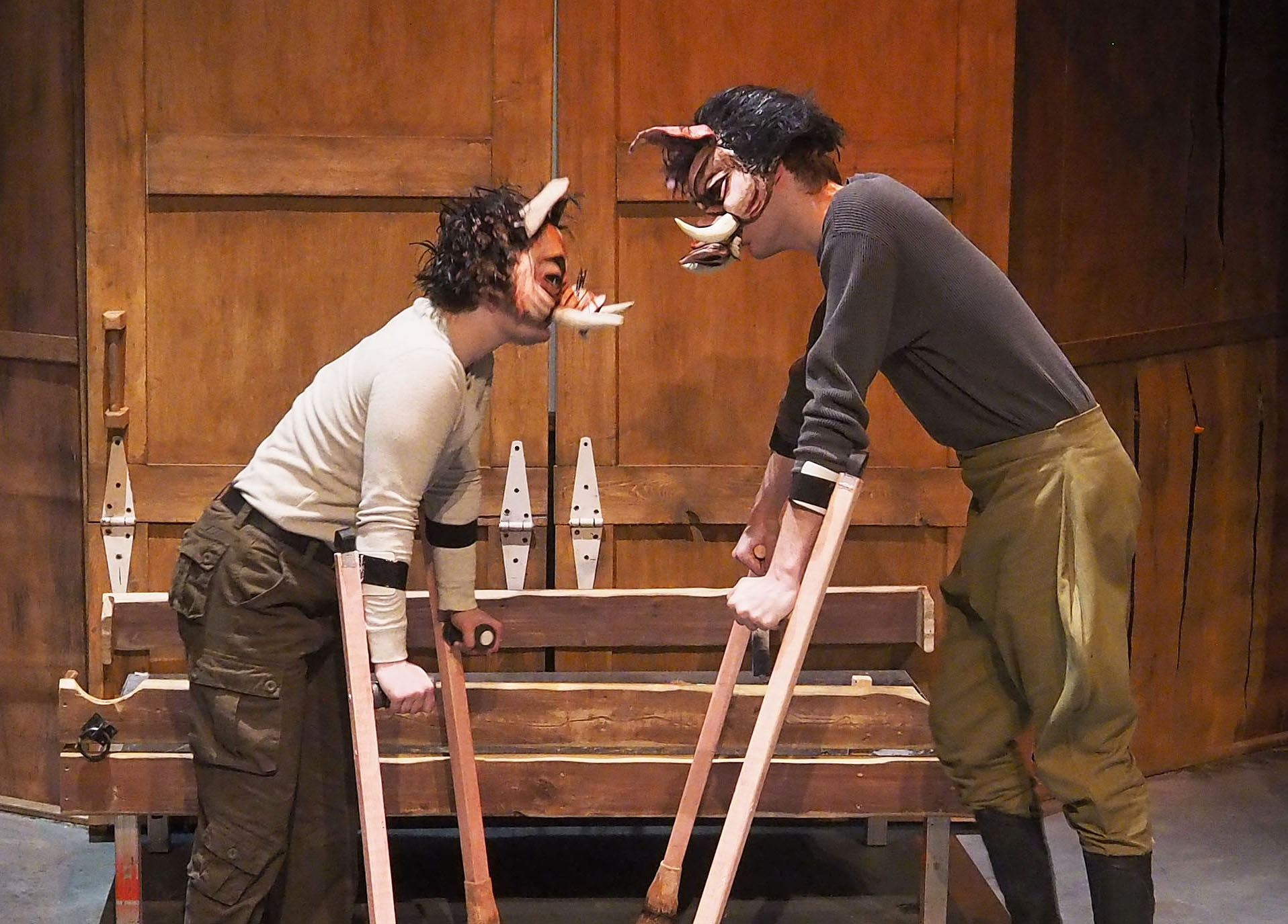 Two performers in animal masks staring at each other. They both lean on crutches. The background is large wooden barn doors. 