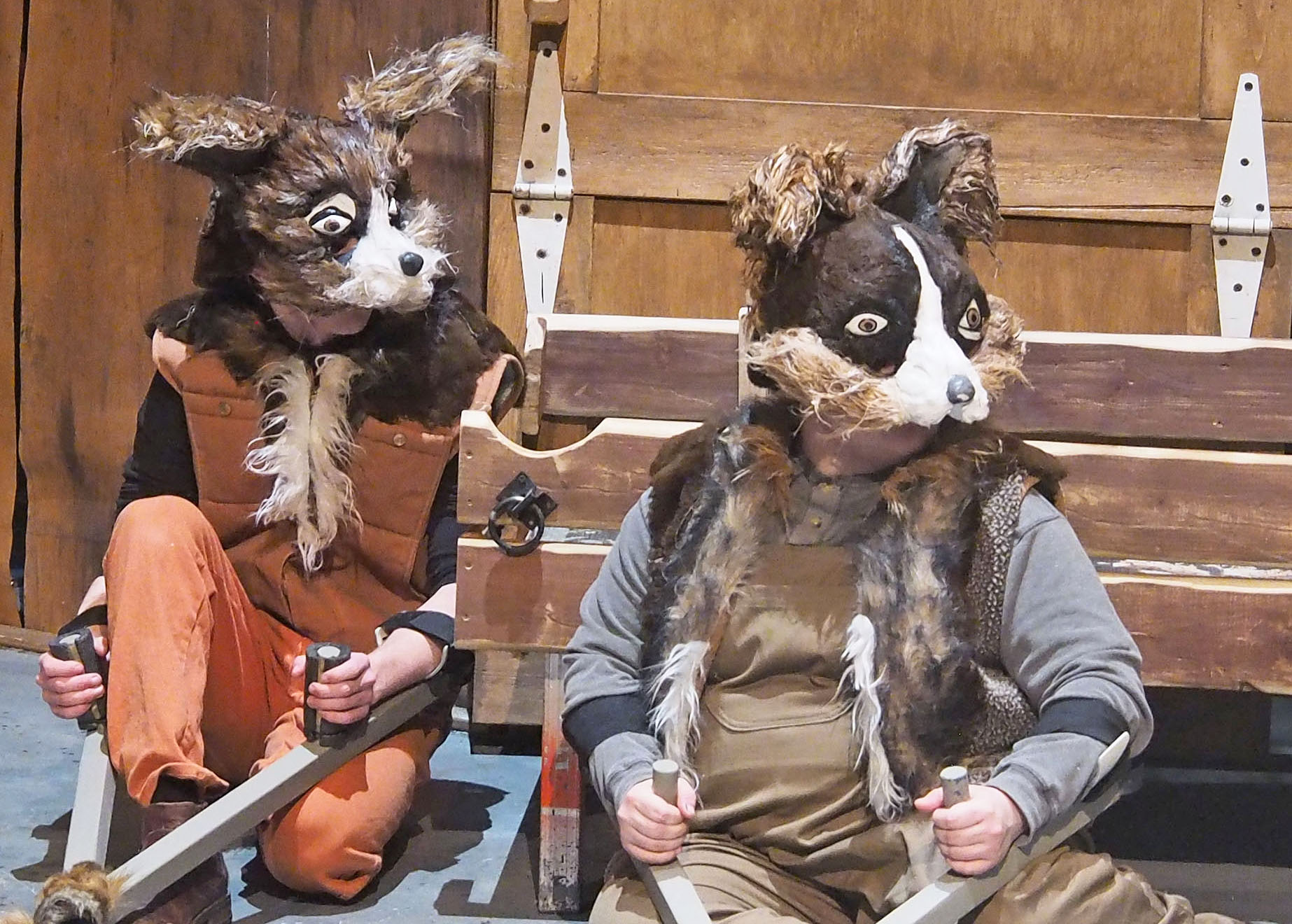 Two performers dressed in animal masks leaning against large barn doors. They look outward. 