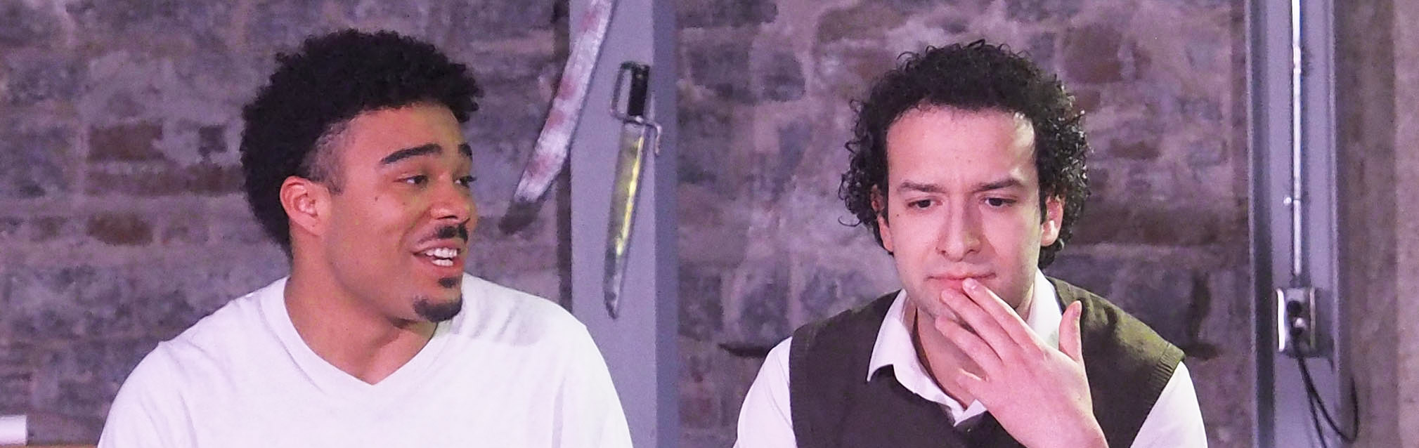 Two performers sit on a brown couch against a brick wall with knives attached to it. One looks contemplatively concerned while the other smiles.