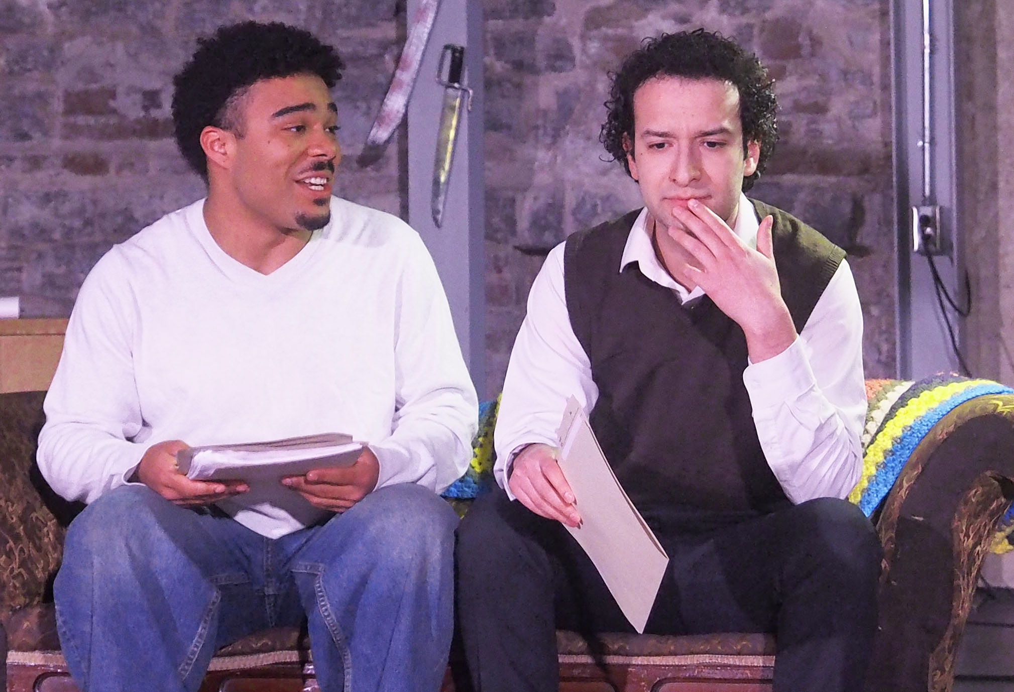 Two performers sit on a brown couch against a brick wall with knives attached to it. They each hold papers. One looks contemplatively concerned while the other smiles. 