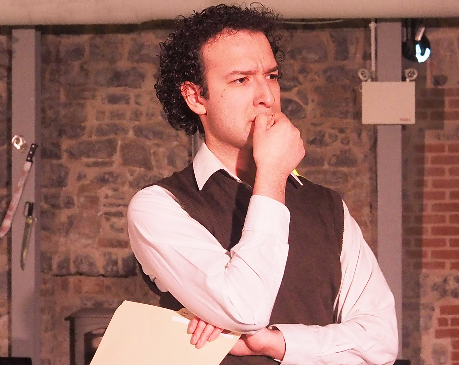 A performer stares contemplatively outward. He holds a paper in a crossed arm and his other hand is at his chin.