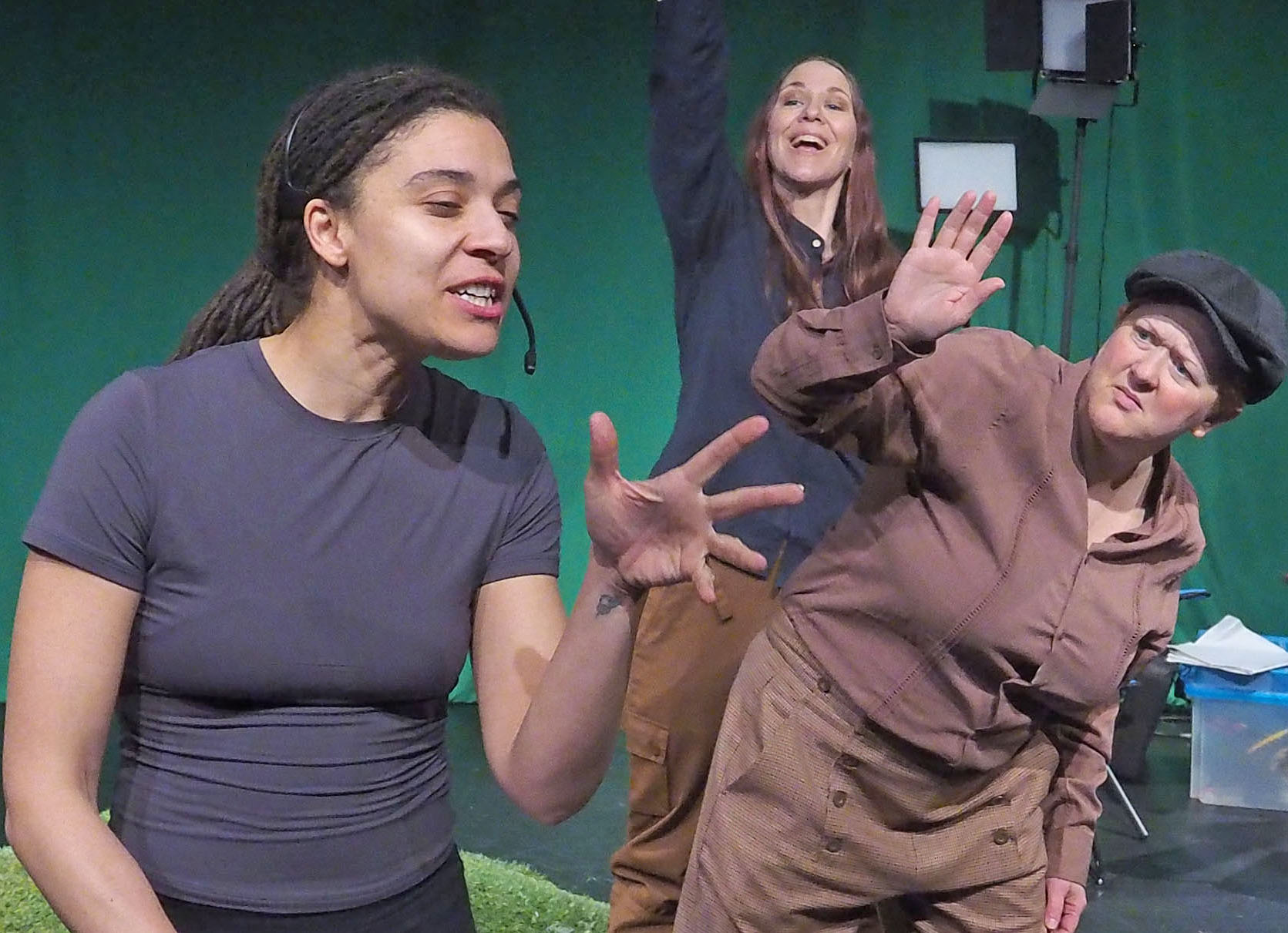 Three performers on stage. On wears a headset and seems to be ignoring the other two, who are desperately trying to get their attention. A green screen and stage lights are in the background. 