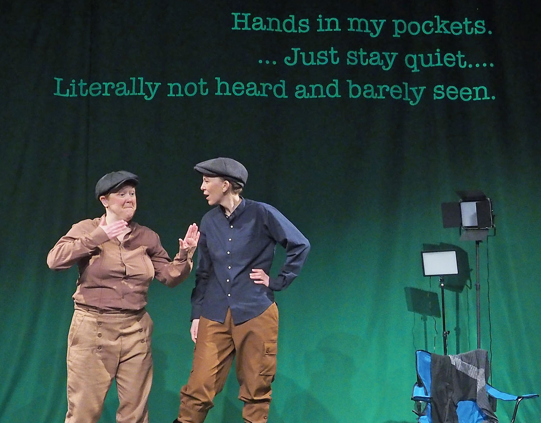 Two performers on stage, dressed in button up shirts and flat cap hats. There are stage lights behind them and the following words appear on a green screen. "Hands in my pockets... Just stay quiet... Literally not heard and barely seen."