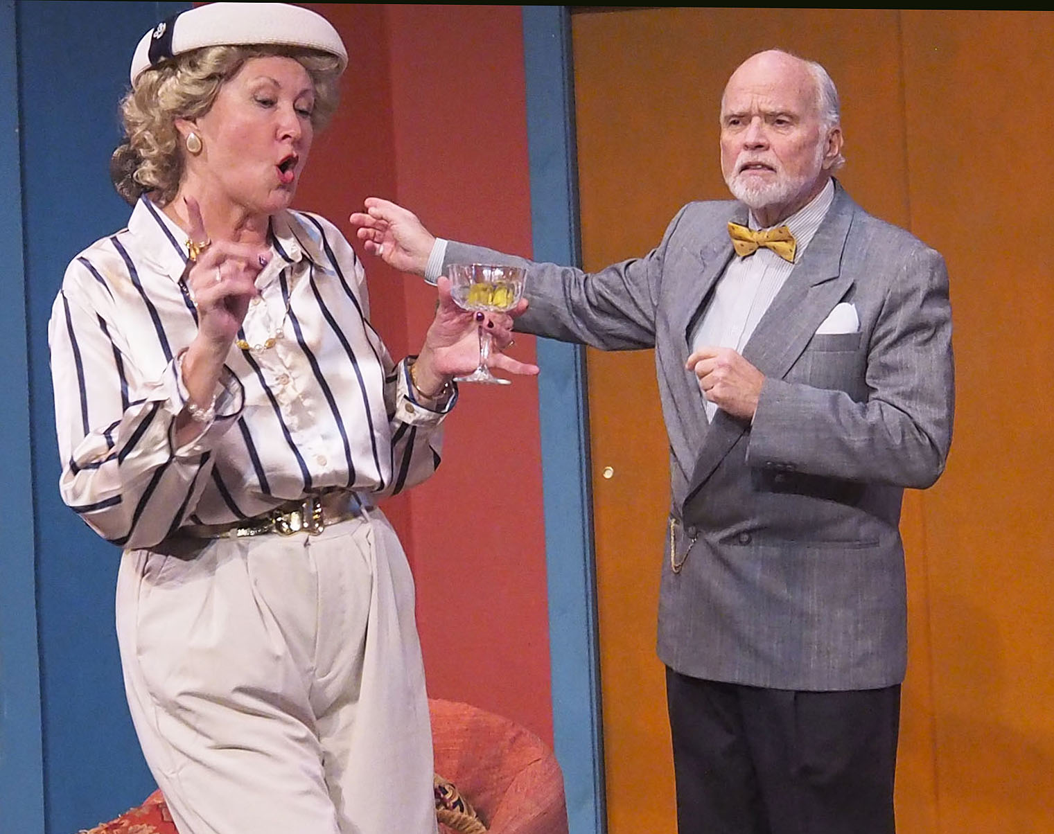 A fashionably dressed woman holds a martini while a man in a suit looks worried and motions his arms outward.